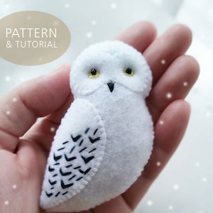 Op de afbeelding: Een witte vilt uil met zwarte geborduurde details. De uil heeft gele ogen en een zwarte snavel. De uil zit in een hand tegen een witte achtergrond met witte stippen.