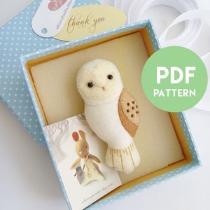 Puede incluir: Un búho de fieltro blanco y marrón con ojos negros y detalles de cuentas marrones en una caja de regalo de lunares azules y blancos. La caja también incluye una tarjeta pequeña con una ilustración de conejo y el texto "whimsylandia.etsy.com". El texto "PDF PATTERN" está en un círculo verde.