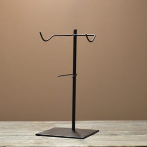 May include: A black metal display stand. It has a square base, a central vertical pole, two curved arms at the top, and a small horizontal bar below. The stand is designed for displaying items.