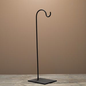 May include: A black metal plant hook stand with a square base and a curved hook at the top. The stand is tall and slender, designed to hold hanging plants or decorative items. The hook extends approximately 12 inches from the top.