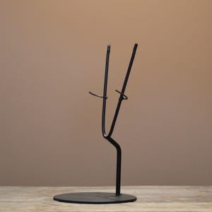 May include: A matte black metal display stand featuring a circular base and a vertical support arm that branches into a V-shaped cradle. The structure is designed with two small, curved hooks extending from the upper arms to hold items securely in place.