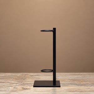 May include: A matte black metal display stand featuring a vertical rectangular post attached to a flat square base, with two circular rings extending horizontally from the post to hold items in a stacked or upright position.