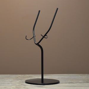 May include: A black metal display stand with a circular base and a curved, abstract design. The stand features two upward-curving arms and a central support, suitable for holding items. The stand is made of metal and has a matte finish.