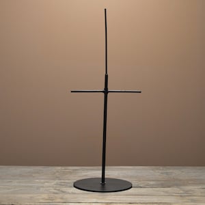 May include: A black metal display stand with a circular base and a vertical pole. The pole has a horizontal crossbar and a smaller vertical extension. The stand is designed to hold items for display.