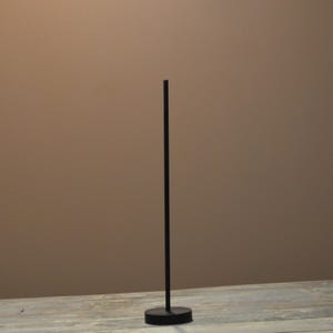 May include: A tall, thin, black metal rod stand featuring a circular base. The vertical rod is centered on the round, flat base, creating a simple, upright structure. The object is finished in a matte black color throughout its entire surface.