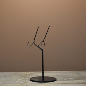 May include: A black metal display stand with a circular base and a curved top. The stand has a minimalist design, with a slender stem and two curved arms extending upwards. The stand is designed to hold an item.