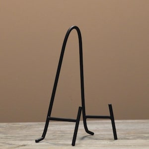 May include: A black metal display stand with a curved top and a three-legged base. The stand is designed to hold plates, artwork, or other decorative items. The stand is made of metal and has a matte finish.