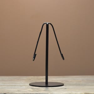 May include: A black metal display stand with a circular base and a vertical support that curves upward, ending in two hooks. The stand is designed to hold an item, likely jewelry or a small decorative object.