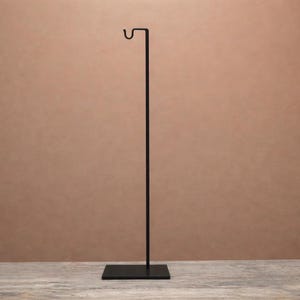 May include: A tall, slender black metal display stand featuring a square base and a single vertical rod that terminates in a curved hook at the top. The structure is composed of a flat, solid square foundation supporting a thin, straight upright pole.