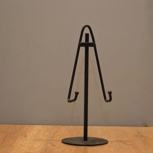 Pode incluir: Suporte de exibição de metal preto com base triangular e dois ganchos. O suporte é projetado para segurar pratos ou outros objetos planos.