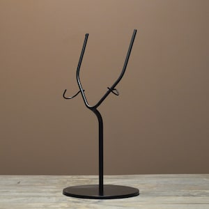 May include: A black metal display stand featuring a circular base, a central vertical rod, and two upward-curving arms with small hooks on each side. The structure is designed to hold items securely in an upright position with a smooth finish.