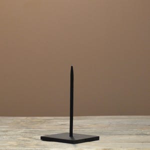 May include: A black metal display stand with a square base and a vertical rod. The stand is designed to hold and showcase an item. The background is a solid brown color, and the stand is placed on a wooden surface.