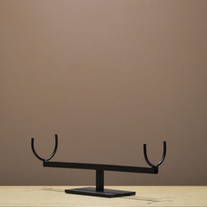 May include: A black metal decorative sculpture with a seesaw design. The sculpture has a rectangular base, a vertical support, a horizontal bar, and curved, U-shaped ends. The piece is set against a neutral background.