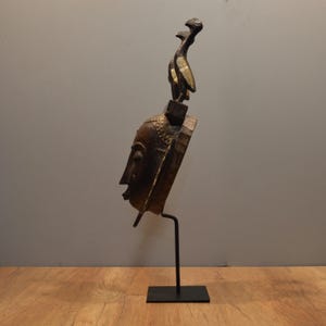 May include: A wooden mask with a human face and two bird figures on top. The mask is painted in gold and brown colors and is displayed on a black metal stand.