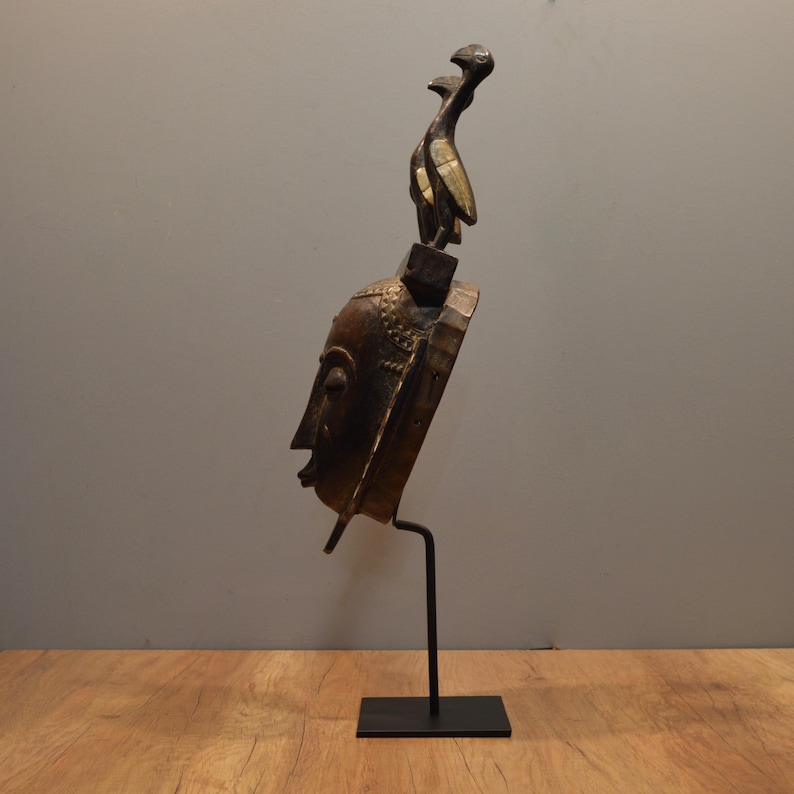 May include: A wooden mask with a human face and two bird figures on top. The mask is painted in gold and brown colors and is displayed on a black metal stand.