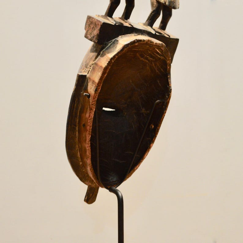 May include: A wooden mask with a carved human face and a stand. The mask is brown and black with a textured surface. The stand is black metal.