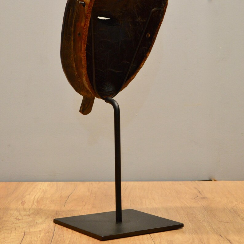 May include: A wooden mask with a dark brown patina, mounted on a black metal stand. The mask has a rounded shape and is carved with a simple design.