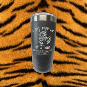Puede incluir: Un vaso de acero inoxidable negro con un gráfico de tigre y el texto "I got peed on by a tiger, property of the tiger king, est. 1999".