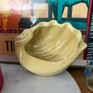 May include: A pale yellow ceramic bowl shaped like a seashell with a glossy finish and a scalloped, wavy rim. The interior of the bowl features curved, ribbed textures that mimic the natural appearance of a clam shell, creating a deep, open basin.