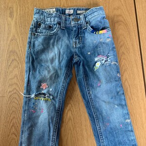 May include: Blue denim jeans with paint splatters and floral embroidery. The jeans have distressed areas and a small pocket on the right side.