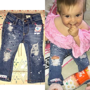 May include: A pair of blue denim jeans with white paint splatters and distressed details. The jeans have a small embroidered patch on the right pocket. The jeans are shown on a child.
