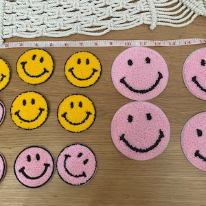 May include: A collection of 10 smiley face patches, 5 yellow and 5 pink. The patches are made of fuzzy fabric and have black embroidered eyes and mouths.