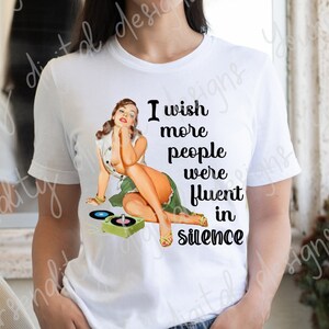 May include: White t-shirt with a retro pin-up girl graphic and the text "I wish more people were fluent in silence." The graphic features a woman in a green skirt and yellow heels with a record player. The shirt is paired with denim shorts.
