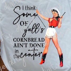 Puede incluir: Camiseta blanca con un diseño gráfico en blanco y negro que presenta a una chica pin-up con un atuendo rojo y blanco sosteniendo un lazo. El texto en la camiseta dice "I think some of yall's cornbread ain't done in the center."