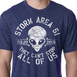 May include: Navy blue t-shirt with a white graphic of an alien head and the text "STORM AREA 51 09.20 2019 THEY CAN'T STOP ALL OF US". The shirt has short sleeves and a crew neck.