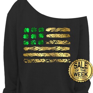 May include: Black off-the-shoulder sweatshirt with a gold and green graphic design. The design resembles a flag with gold stripes and green shamrocks. A gold sticker on the right side says "SALE OF THE WEEK ENDS SUNDAY."