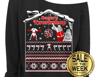 May include: Black off-the-shoulder sweatshirt with a festive graphic. The design features a house-shaped illustration with the text "Santa's Twerkshop", Santa Claus, a disco ball, and twerking figures. A gold "Sale of the Week" sticker is visible.