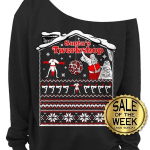 May include: Black off-the-shoulder sweatshirt with a festive graphic. The design features a house-shaped illustration with the text "Santa's Twerkshop", Santa Claus, a disco ball, and twerking figures. A gold "Sale of the Week" sticker is visible.