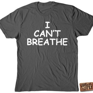 May include: Dark gray t-shirt with the white text "I CAN'T BREATHE" in a bold, sans-serif font. The shirt is a crew neck style and appears to be made of a soft material. A small, rectangular logo is visible in the bottom right corner.
