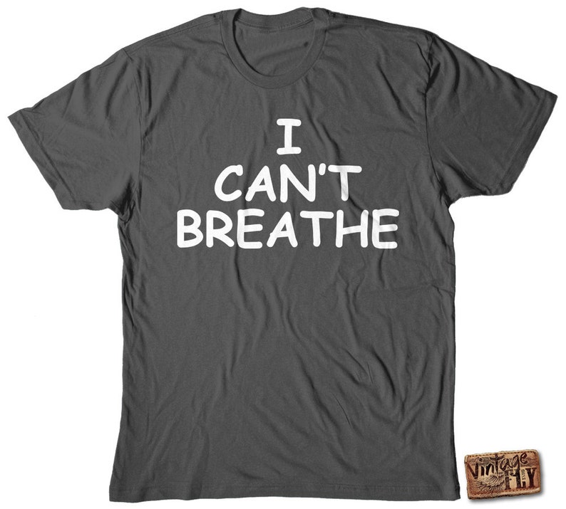 May include: Dark gray t-shirt with the white text "I CAN'T BREATHE" in a bold, sans-serif font. The shirt is a crew neck style and appears to be made of a soft material. A small, rectangular logo is visible in the bottom right corner.