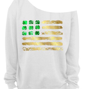 May include: White off-the-shoulder sweatshirt featuring a gold and green design. The design resembles the American flag, with green shamrocks replacing the stars and gold stripes. The sweatshirt has a relaxed fit and long sleeves.