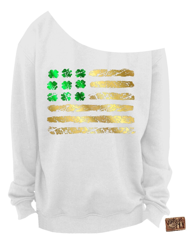 May include: White off-the-shoulder sweatshirt featuring a gold and green design. The design resembles the American flag, with green shamrocks replacing the stars and gold stripes. The sweatshirt has a relaxed fit and long sleeves.