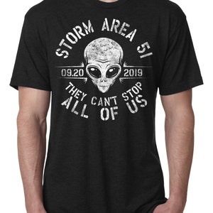May include: Dark gray t-shirt with a white graphic of an alien head and the text "STORM AREA 51 09.20.2019 THEY CAN'T STOP ALL OF US". The shirt has short sleeves and a crew neck.