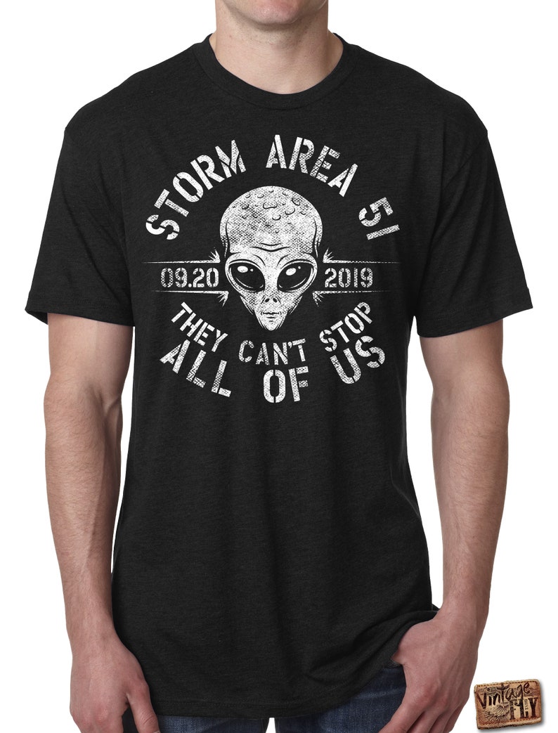 May include: Dark gray t-shirt with a white graphic of an alien head and the text "STORM AREA 51 09.20.2019 THEY CAN'T STOP ALL OF US". The shirt has short sleeves and a crew neck.