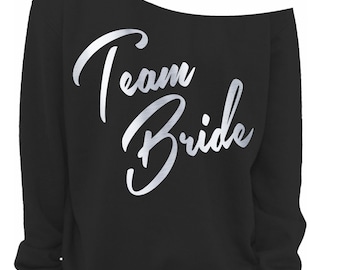 May include: Black off-the-shoulder sweatshirt with the words "Team Bride" in a silver, cursive font. The sweatshirt has long sleeves and a relaxed fit. A small, rectangular logo is visible in the bottom right corner.