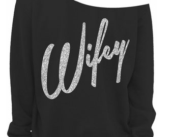 May include: Black off-the-shoulder sweatshirt featuring the word "Wifey" in silver glitter script. The top has long sleeves and a relaxed fit, suitable for everyday wear. A stylish and comfortable garment.