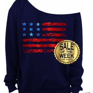 May include: Navy blue off-the-shoulder sweatshirt featuring a distressed American flag design in red and blue. The design includes nine blue stars and red stripes. A gold seal with the text "SALE OF THE WEEK ENDS SUNDAY" is also visible.