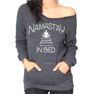 May include: Dark gray off-the-shoulder sweatshirt with the white text "NAMAST'AY IN BED" and a graphic of a person meditating on a bed. The sweatshirt has long sleeves and a front pocket.