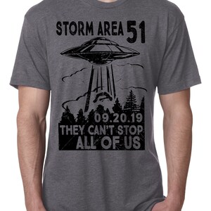 May include: Gray t-shirt with a black graphic of a UFO beaming down on a forest. The text reads "STORM AREA 51 09.20.19 THEY CAN'T STOP ALL OF US". The shirt is short-sleeved and has a crew neck.
