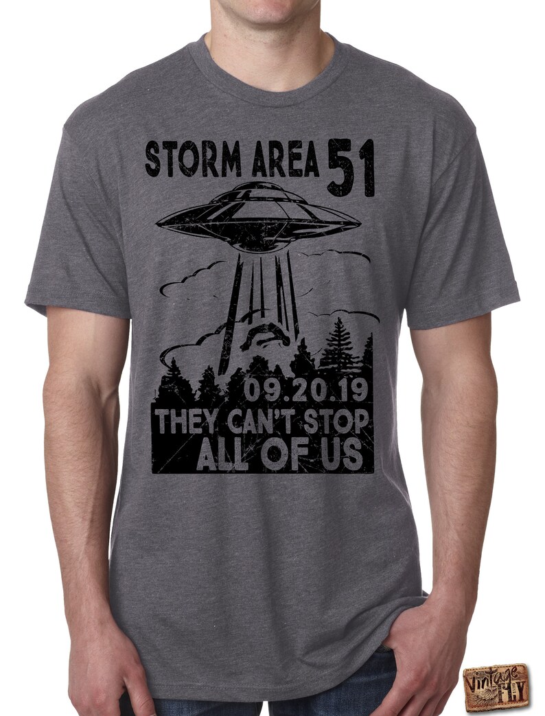 May include: Gray t-shirt with a black graphic of a UFO beaming down on a forest. The text reads "STORM AREA 51 09.20.19 THEY CAN'T STOP ALL OF US". The shirt is short-sleeved and has a crew neck.