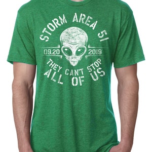May include: Green t-shirt with a white graphic featuring an alien head and the text "STORM AREA 51 09.20 2019 THEY CAN'T STOP ALL OF US". The shirt has short sleeves and a crew neck.