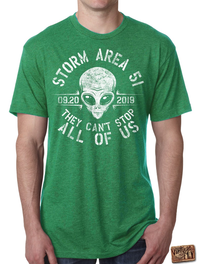 May include: Green t-shirt with a white graphic featuring an alien head and the text "STORM AREA 51 09.20 2019 THEY CAN'T STOP ALL OF US". The shirt has short sleeves and a crew neck.