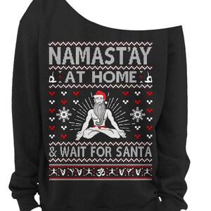 May include: Black off-the-shoulder sweatshirt with a festive design. The text reads "NAMAST'AY AT HOME & WAIT FOR SANTA" above an illustration of a meditating Santa Claus. The design includes snowflakes and yoga poses.