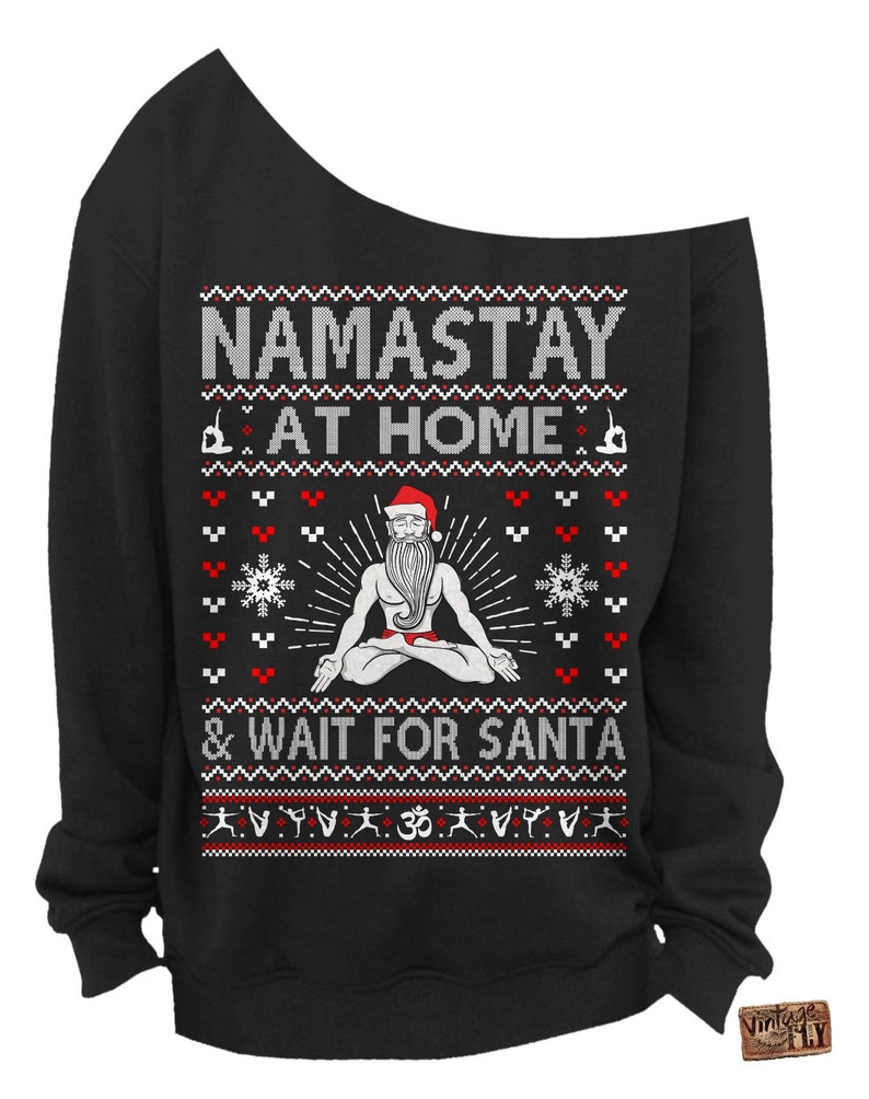May include: Black off-the-shoulder sweatshirt with a festive design. The text reads "NAMAST'AY AT HOME & WAIT FOR SANTA" above an illustration of a meditating Santa Claus. The design includes snowflakes and yoga poses.