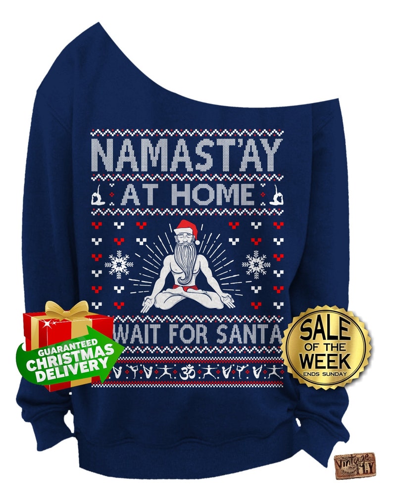 May include: Navy blue off-the-shoulder sweatshirt with a festive design. The text "NAMAST'AY AT HOME" and "WAIT FOR SANTA" are displayed. The design includes a Santa Claus figure in a yoga pose, snowflakes, and yoga poses.