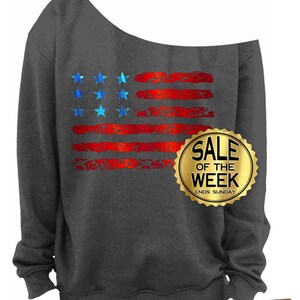May include: Dark gray off-the-shoulder sweatshirt featuring a distressed American flag design in red and blue. The sweatshirt has the text "SALE OF THE WEEK ENDS SUNDAY" on a gold seal.
